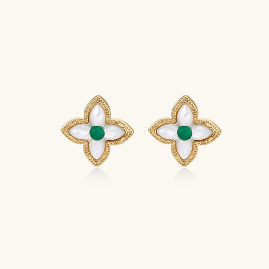 Gold quatrefoil stud earrings with white mother-of-pearl petals and emerald green gemstone center on beige background.